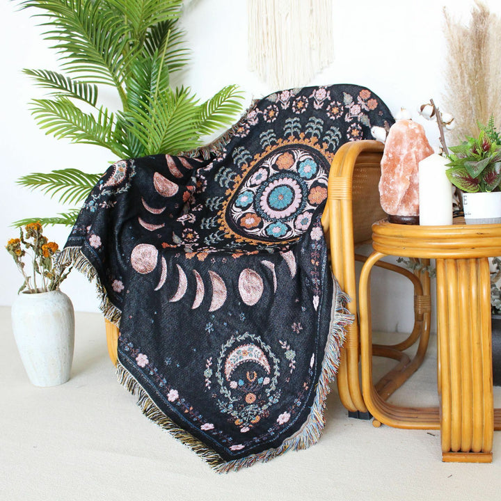 Floral Phases of the Moon Woven Blanket draped over a chair, featuring celestial and floral patterns. Made from 100% combed cotton, this versatile piece adds a serene, mystical vibe to your space.