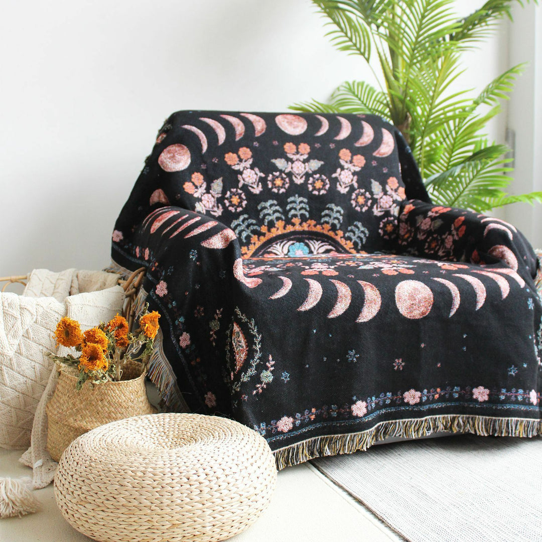 Floral Phases of the Moon Woven Blanket draped over a sofa chair, featuring celestial and floral patterns. Made from 100% combed cotton, this versatile piece adds a mystical touch to any space.