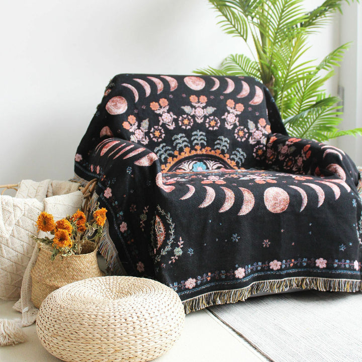 Floral Phases of the Moon Woven Blanket draped over a sofa chair, featuring celestial and floral patterns. Made from 100% combed cotton, this versatile piece adds a mystical touch to any space.