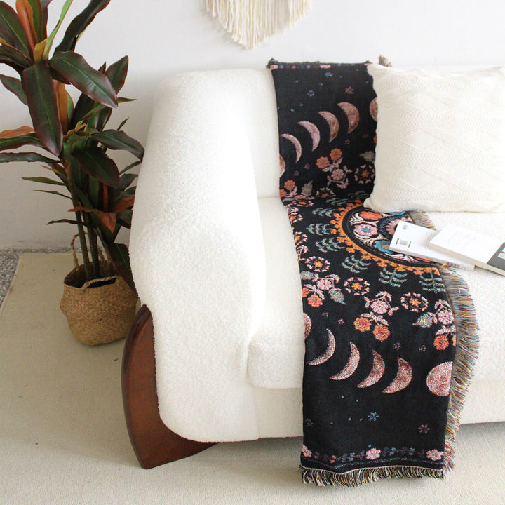 Floral Phases of the Moon Woven Blanket folded on a couch, featuring celestial and floral patterns. Made from 100% combed cotton, this versatile blanket adds a serene, mystical touch to your space.