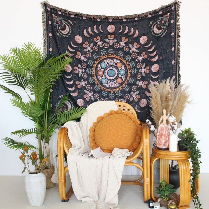 Floral Phases of the Moon Woven Blanket hanging on the wall behind a chair and plants, featuring celestial and floral patterns. Made from 100% combed cotton, this versatile piece adds a mystical touch to your space.