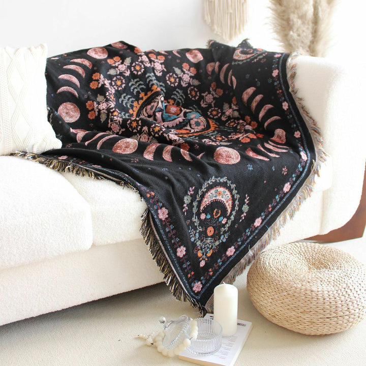 Floral Phases of the Moon Woven Blanket thrown on a couch, showcasing its celestial and floral patterns. Made from 100% combed cotton, this versatile blanket adds a mystical and cozy touch to your space.