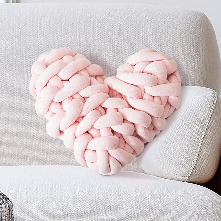 VibeCore Heart-Shaped Knot Throw Pillow in baby pink, placed on a couch, featuring its heart-shaped, woven knot design. This plush, cozy pillow adds a charming and loving touch to your living space, creating a warm, inviting atmosphere.