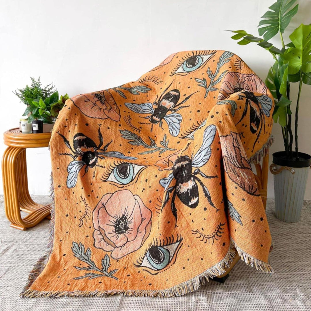 VibeCore Honey Dreams Woven Blanket draped over chair, featuring a detailed bee motif and captivating eye patterns. Made from soft, breathable 100% combed cotton, it adds warmth, charm, and a unique touch to your space with its vibrant, reversible design.