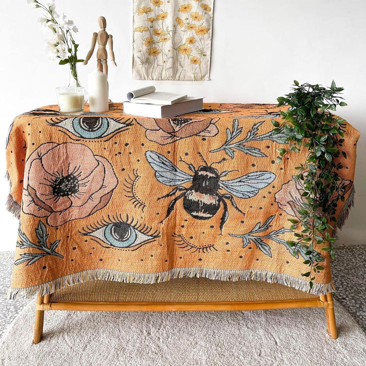 VibeCore Honey Dreams Woven Blanket on table, featuring a detailed bee motif and captivating eye patterns. Crafted from soft, breathable 100% combed cotton, it adds a charming and unique touch to your space, elevating any surface with its vibrant, reversible design.