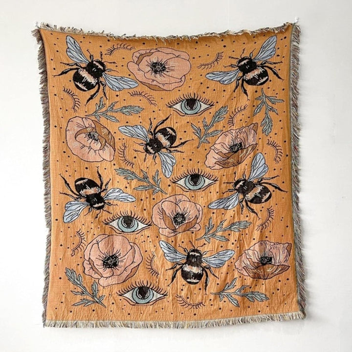 VibeCore Honey Dreams Woven Blanket product view, showcasing a detailed bee motif and captivating eye patterns. Crafted from soft, breathable 100% combed cotton, it offers two reversible designs for versatile styling, adding warmth, charm, and a unique vibe to any room.