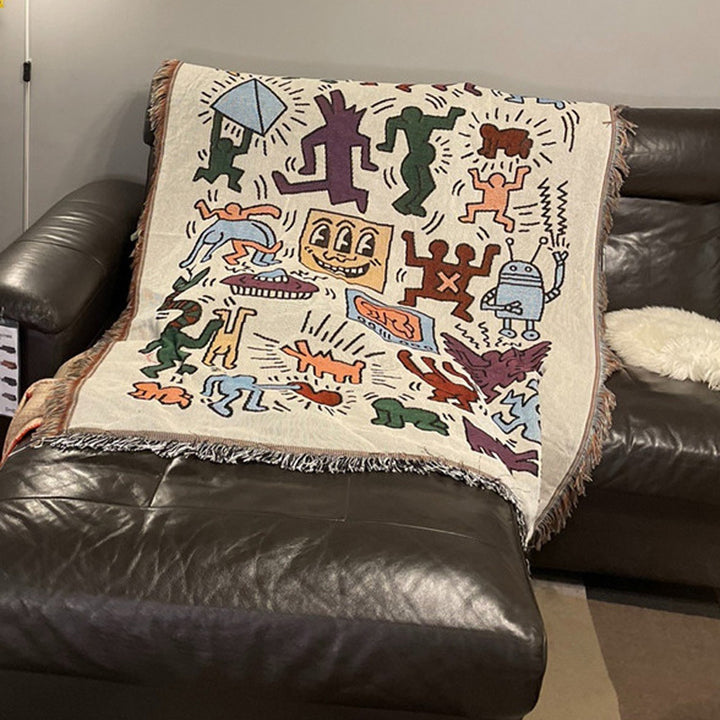 VibeCore Keith Haring Graffiti Woven Blanket completely splayed out on the corner of couch, featuring bold street art design and vibrant 100% cotton fabric.