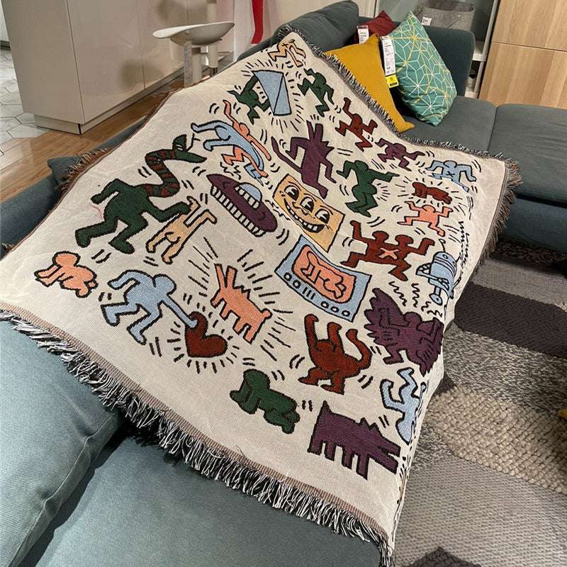 VibeCore Keith Haring Graffiti Woven Blanket on blue couch, featuring bold street art design and vibrant 100% cotton fabric.