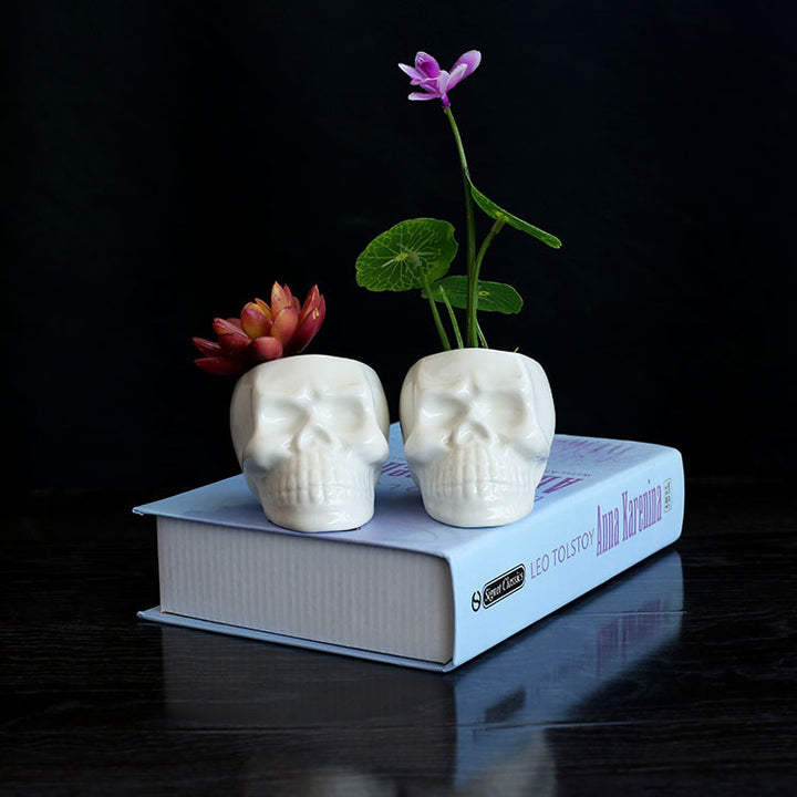 Two mini skull planters on a book, showcasing small plants in an edgy, ceramic design.