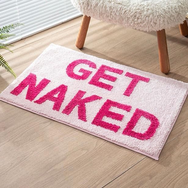 Aesthetic "Pink Get Naked Bath Mat" displayed in front of a chair, featuring a vibrant pink color and playful design. The soft, plush texture adds comfort and personality to the space, while the non-slip backing ensures stability and safety, making it a fun and functional addition to any room.