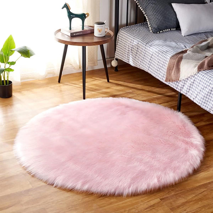 The VibeCore Round Faux Fur Accent Rug in Light Pink placed beside a bed, showcasing its soft, plush texture and chic round shape, adding a cozy and stylish touch to the room's decor.