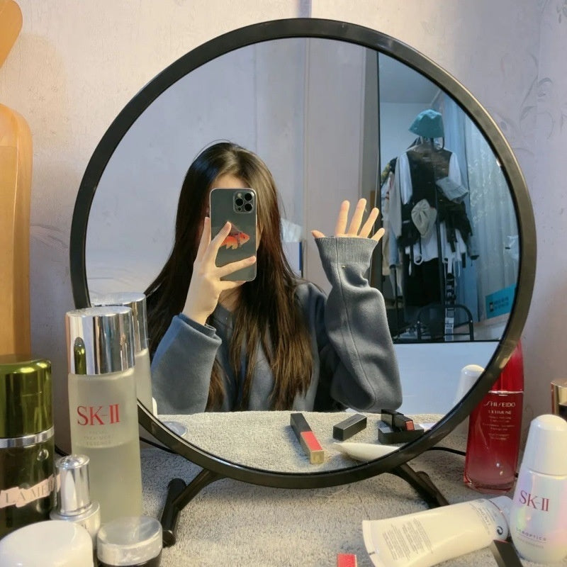 A black round vanity mirror featuring a sleek and minimalist design, reflecting a girl taking a selfie. The mirror offers a high-resolution reflection, enhancing beauty routines with a flawless view.