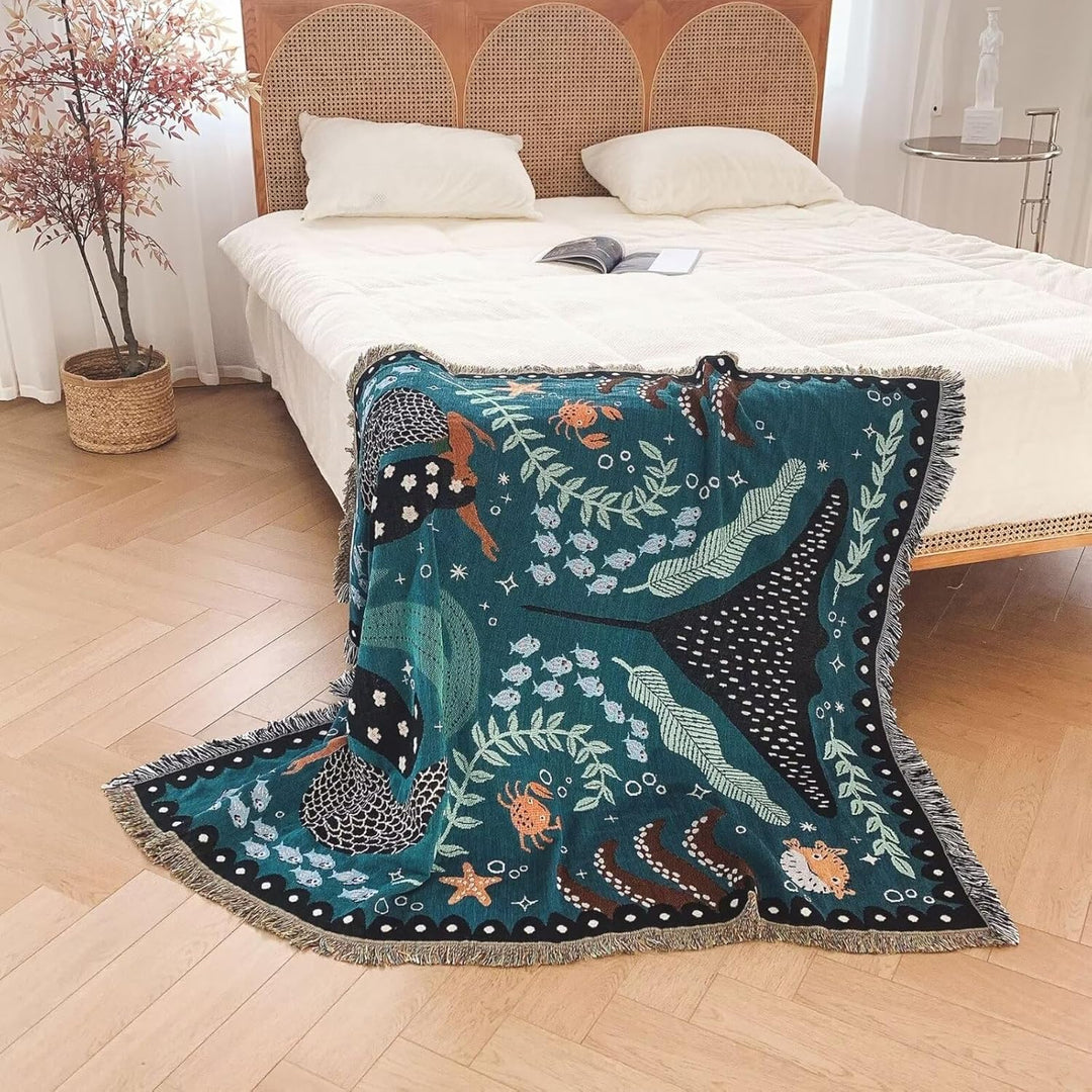 VibeCore Swimming Mermaids Woven Blanket draped over the corner of bed, showcasing an enchanting mermaid design and soft 100% combed cotton fabric.