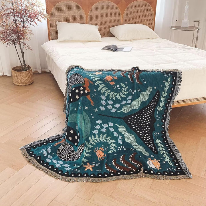 VibeCore Swimming Mermaids Woven Blanket draped over the corner of bed, showcasing an enchanting mermaid design and soft 100% combed cotton fabric.