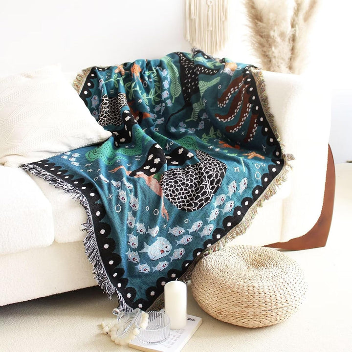 VibeCore Swimming Mermaids Woven Blanket draped over couch, showcasing enchanting mermaid design and vibrant reversible patterns in soft 100% combed cotton.