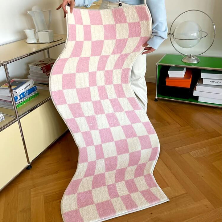Aesthetic pink "Wavy Checkerboard Accent Rug" being held up, showcasing its dynamic checkerboard pattern with a wavy twist. This close-up highlights the soft, plush texture and non-slip backing, perfect for adding a playful touch and comfort to any room.