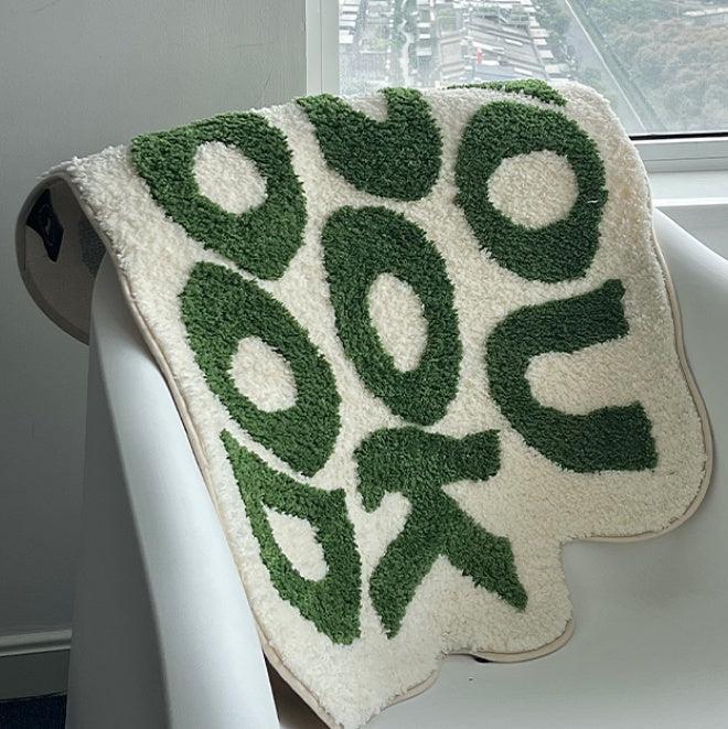 VibeCore You Look Good Accent Rug in green, folded over a chair, showcasing its uplifting quote design, soft plush texture, and non-slip backing. This cute rug adds a playful and modern touch to bedrooms, dorms, or living rooms. Its vibrant, motivational design brings personality and comfort to any space, perfect for contemporary decor. A cozy, fun accent rug that complements your home with its chic and cheerful style. Ideal for those looking for a fun, aesthetic rug to elevate their room.