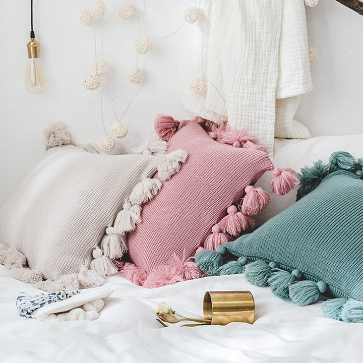 VibeCore Nordic Style Hand-Knitted Pillow Covers in Blossom, Dark Gray, and Ivory arranged on a bed, featuring boho fringe details and a cozy, textured look.