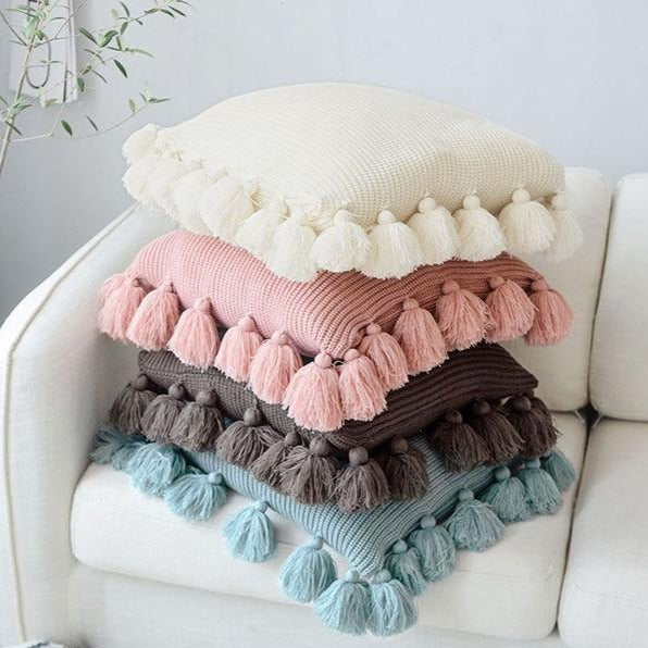 VibeCore Nordic Style Hand-Knitted Pillow Covers in 4 colors (blossom, ivory, dark gray, turquoise blue) stacked together in a cozy living room, showcasing fringe details for a boho-inspired touch.